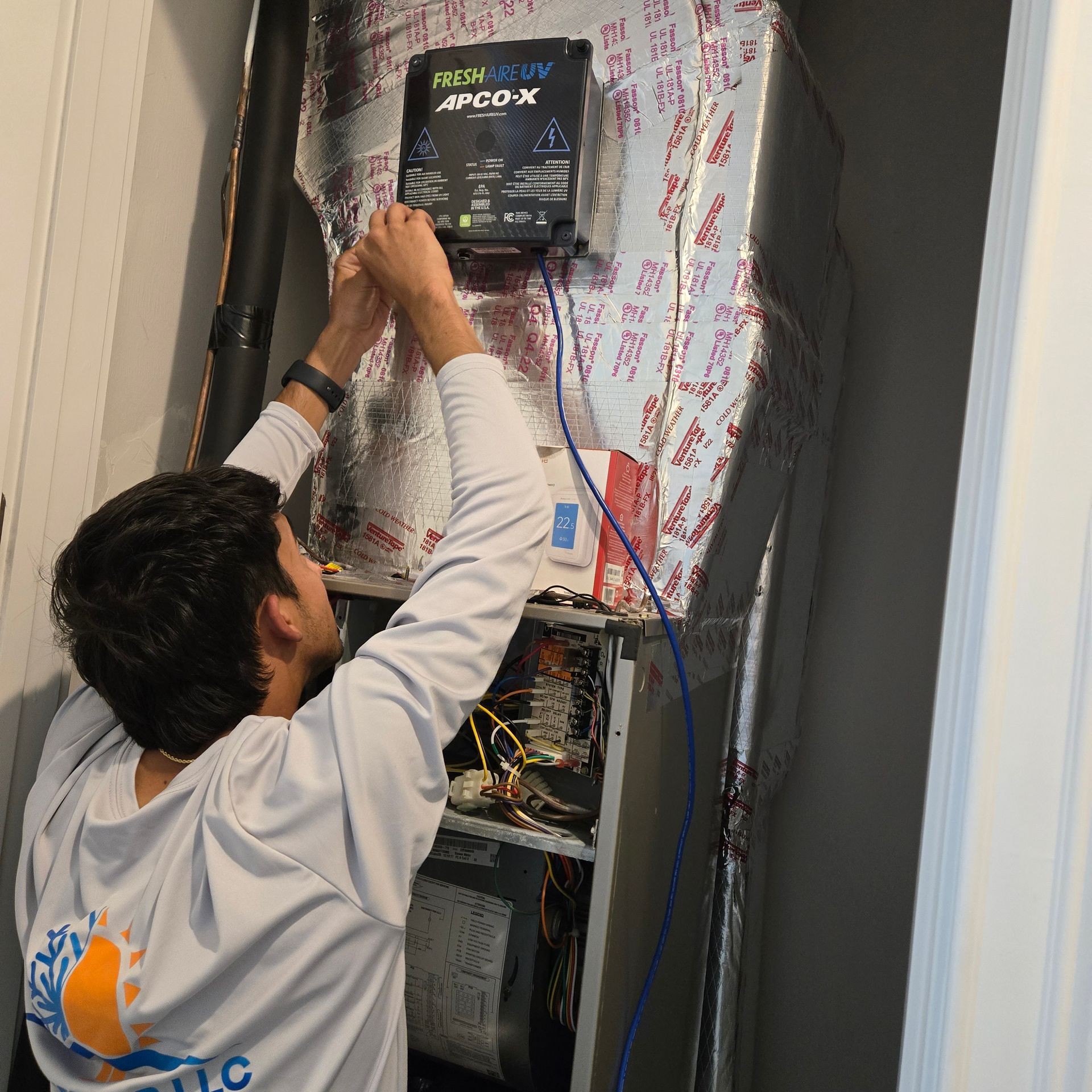 Technician installing APCO-X air purifier on an HVAC system with reflective insulation.